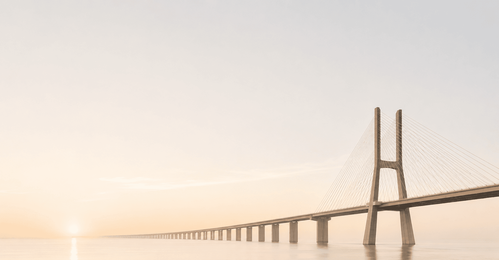 A long cable-stayed bridge reaching toward the horizon at sunrise over calm water, beneath a vast, soft, warm sky.